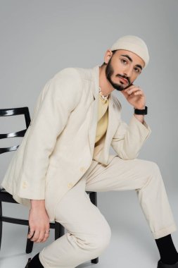 Gri arka planda sandalyede poz veren şapkalı ve bej takım elbiseli moda homoseksüel adam. 