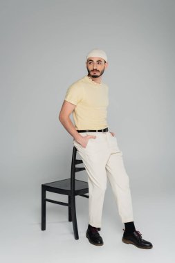 Gri arka planda sandalyeye yakın poz veren bej şapkalı genç eşcinsel adam. 
