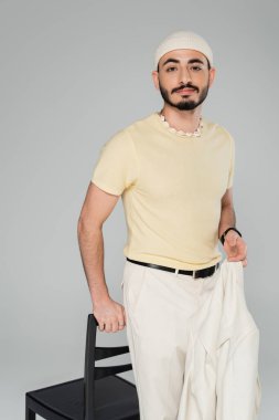 Şapkalı sakallı eşcinsel adam gri sandalyenin yanında bej ceket giyiyor.  