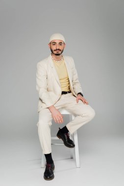 Gri arka planda, sandalyede oturan bej takım elbiseli ve şapkalı, modaya uygun eşcinsel bir adam. 