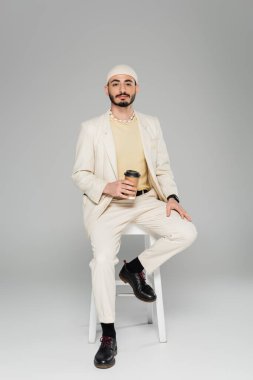 Gri arka planda sandalyede otururken elinde kahve tutan modaya uyan takım elbiseli ve şapkalı bir gay. 