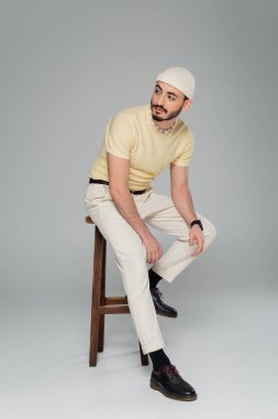 Gri arka planda, sandalyede oturan bej şapkalı, modaya uygun bir gay. 