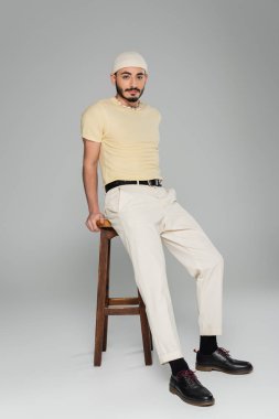 Gri arka planda sandalyenin yanında poz veren şapkalı şık bir gay. 