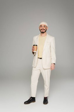 Takım elbiseli neşeli eşcinsel adam yanağında lgbt bayrağı gri arka planda kağıt bardak taşıyor. 