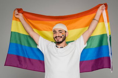 Şapkalı gülümseyen homoseksüel adam gri üzerinde lgbt bayrağı taşıyor.  
