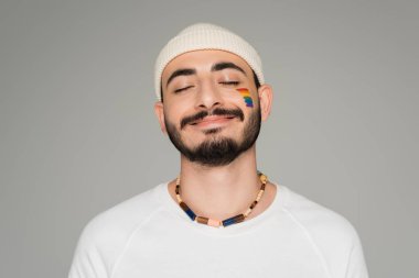 Yanağında lgbt bayrağı olan şapkalı gülümseyen gay adamın portresi. Gri üzerinde tek başına duruyor.  