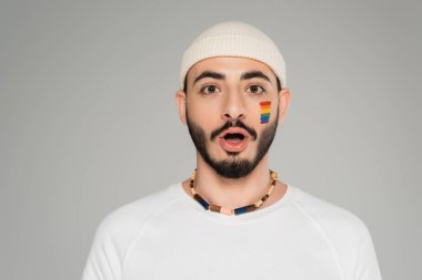 Yanağında lgbt bayrağı olan şapkalı eşcinsel adam gri renkli kameraya bakıyor.  