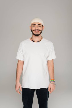 Elinde lgbt bilekliğiyle şapkalı gülümseyen eşcinsel adam gri renkli kameraya bakıyor.  