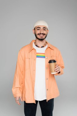 Tişörtünde lgbt bayrağı olan gülümseyen homoseksüel bir adam gri üzerine izole edilmek için elinde kahve tutuyordu.  