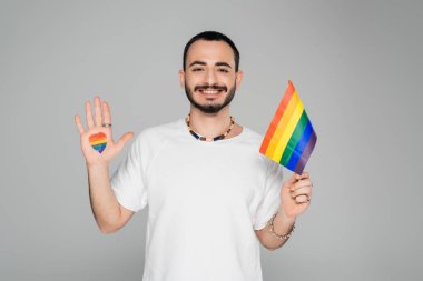 Elinde lgbt bayrağı ve kalbiyle gülümseyen eşcinsel adam homofobiye karşı gri, uluslararası bir günde izole edilmiş kameraya bakıyor.