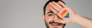Neşeli homoseksüel adam elinde lgbt bayrağıyla yüzünü kapatıyor. Gri pankartta tek başına. 