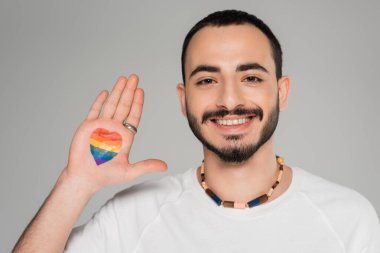 Elinde lgbt bayrağı olan neşeli genç homoseksüel adam gri renkli kameraya bakıyor.  