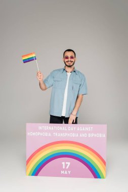 Uluslararası Homofobi Karşıtı Gün, Transfobi ve gri arkaplanda Bifobi harfleri yazan bir pankart yanında lgbt bayrağı tutan neşeli eşcinsel adam. 