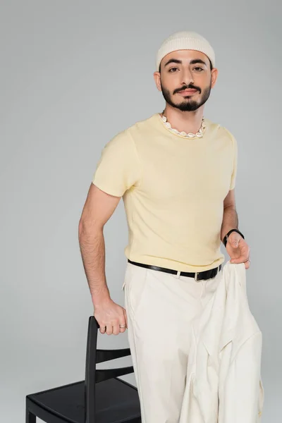 Pria Berjenggot Gay Bertopi Memegang Jaket Krem Dekat Kursi Terisolasi — Stok Foto