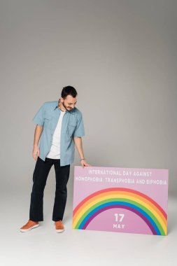 Full length of smiling gay man standing near placard with International Day Against Homophobia, Transphobia and Biphobia lettering on grey  clipart