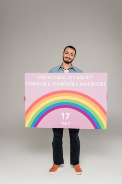 gay man smiling and holding placard with International Day Against Homophobia, Transphobia and Biphobia lettering on grey  clipart