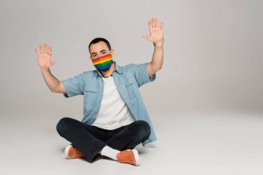 lgbt bayraklı sağlık maskeli esmer eşcinsel adam gri arka planda el sallıyor. 