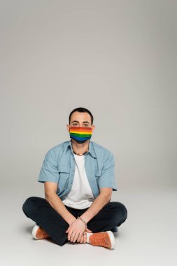 Young homosexual man in medical mask with lgbt flag sitting on grey background  clipart