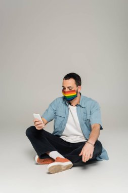 Gri arka planda akıllı telefon kullanan, lgbt bayrağı renginde sağlık maskeli eşcinsel bir adam. 