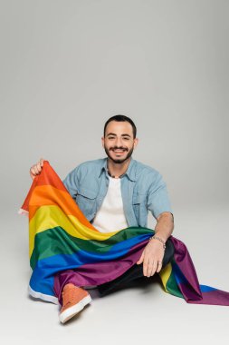 Gri arka planda otururken elinde lgbt bayrağı tutan olumlu eşcinsel adam 