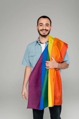 Gri, uluslararası homofobi gününde kameraya gülümseyen lgbt bayrağıyla genç sakallı eşcinsel adam.  