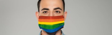 Sağlık maskeli genç esmer adam lgbt bayrağıyla gri pankartta izole edilmiş kameraya bakıyor. 