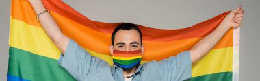 Sağlık maskeli esmer adam gri pankartta LGBT bayrağı taşıyor. 