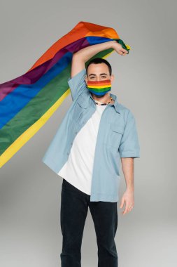 Gri, uluslararası homofobi gününde elinde lgbt bayrağı tutan tıbbi maskeli genç homoseksüel adam.  