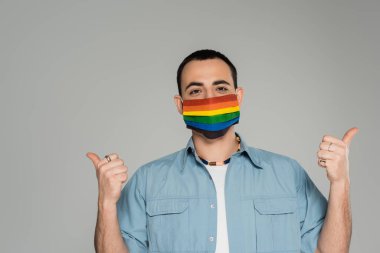 Esmer, sağlık maskeli, lgbt renginde eşcinsel bir adam, homofobiye karşı uluslararası, gri bir günde izole edilmiş bir jest gibi görünüyor.