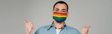 Sağlık maskeli genç homoseksüel adam lgbt bayraklı. Gri pankartta baş parmakları görünüyor. 
