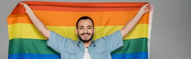 Tasasız homoseksüel adam elinde lgbt bayrağı tutuyordu ve gri pankartta izole edilmiş kameraya bakıyordu. 