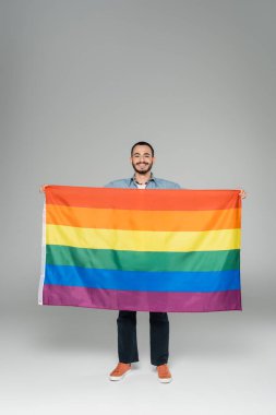 Full length of young gay man holding lgbt flag and smiling at camera on grey background, International Day Against Homophobia clipart