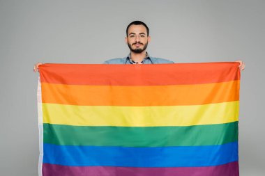 Bearded homosexual man holding lgbt flag and looking at camera isolated on grey   clipart