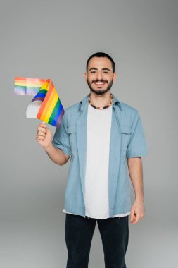 Smiling homosexual man holding lgbt flags and looking at camera isolated on grey   clipart
