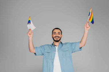 Genç ve neşeli gay adam elinde lgbt bayrakları ve gri kameraya bakıyor.  