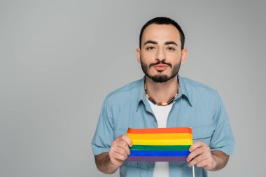 Esmer eşcinsel adam dudaklarını büzüyor ve elinde lgbt bayrağı tutuyor. Gri, Uluslararası Homofobiye Karşı Günü 'nde izole edilmiş. 