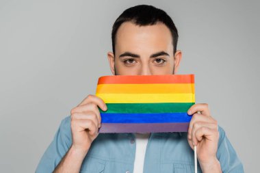 Genç, esmer, eşcinsel bir adam, homofobikliğe karşı, gri, uluslararası bir günde izole edilmiş, lgbt bayrağıyla yüzünü kapatıyor. 