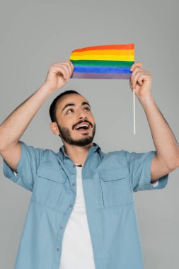 Neşeli ve iyi giyimli gay adam Gri üzerinde izole edilmiş Igbt bayrağına bakıyor  
