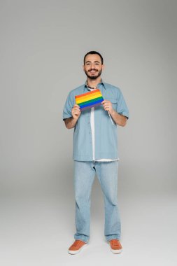 Gri arkaplanda lgbt bayrağı taşıyan kot gömlekli, gülümseyen homoseksüel bir adam. 