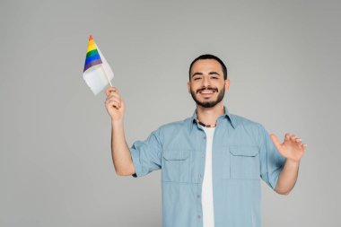 Sakallı, tişörtlü, gülümseyen ve elinde gri renkli lgbt bayrağı tutan eşcinsel adam.  