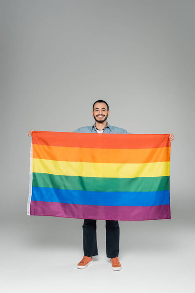 Full length of young gay man holding lgbt flag and smiling at camera on grey background, International Day Against Homophobia