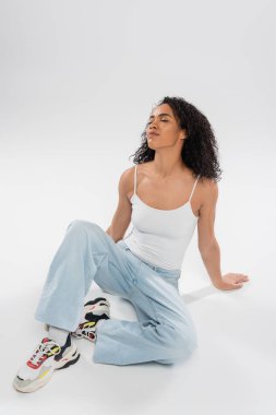 full length of young african american woman in jeans relaxing with closed eyes while sitting on grey background clipart