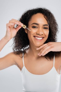 pleased african american woman looking at camera and applying cosmetic serum isolated on grey clipart