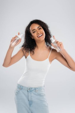 overjoyed african american woman in white tank top posing with bottles of face foam isolated on grey clipart