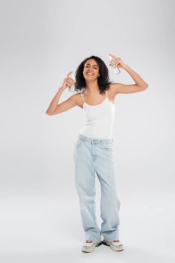 full length of cheerful african american woman in blue jeans posing with bottles of face foam on grey background clipart