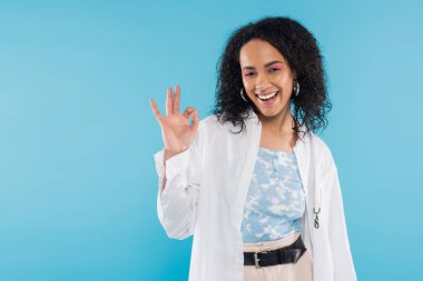 overjoyed african american woman in stylish shirt smiling at camera and showing okay sign isolated on blue clipart