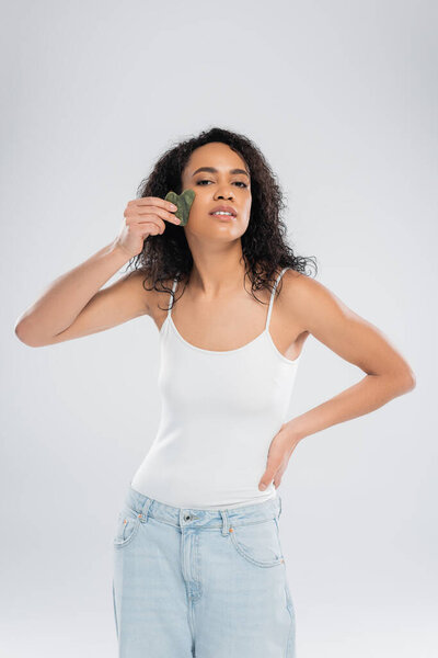 young african american woman doing face massage with stone scraper while posing with hand on hip isolated on grey