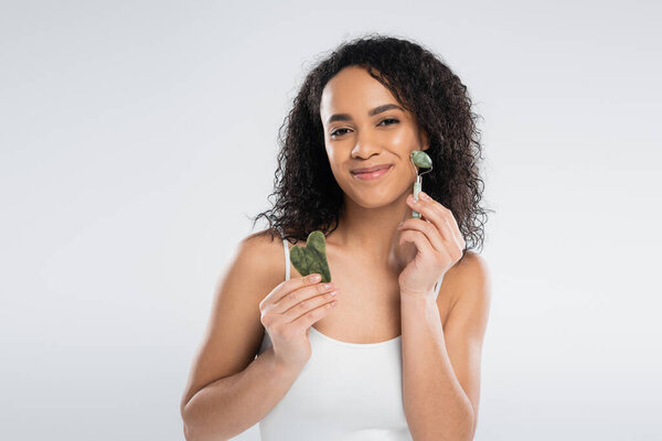 joyful african american woman holding face scraper and using jade roller isolated on grey