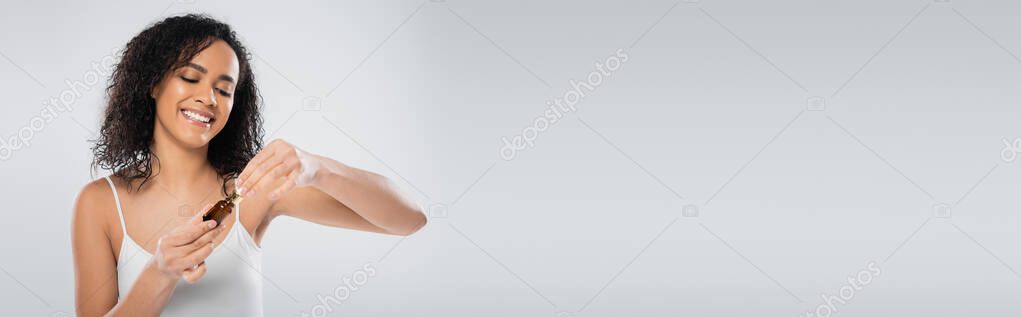Brunette african american woman in tank top opening cosmetic serum isolated on grey, banner