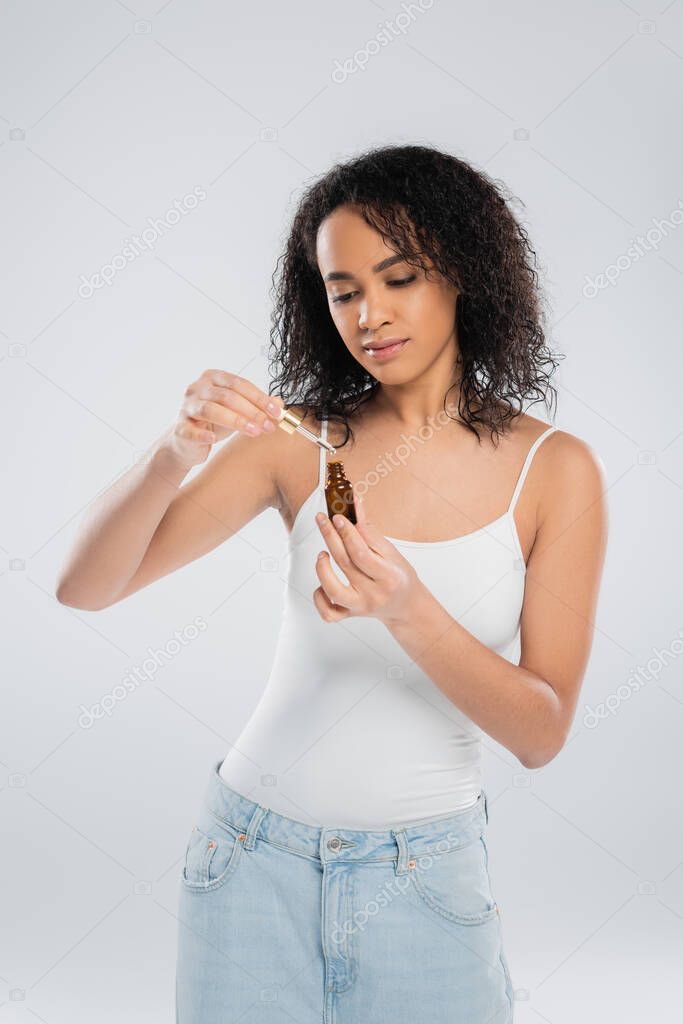 Brunette african american woman holding dropper and bottle of cosmetic serum isolated on grey